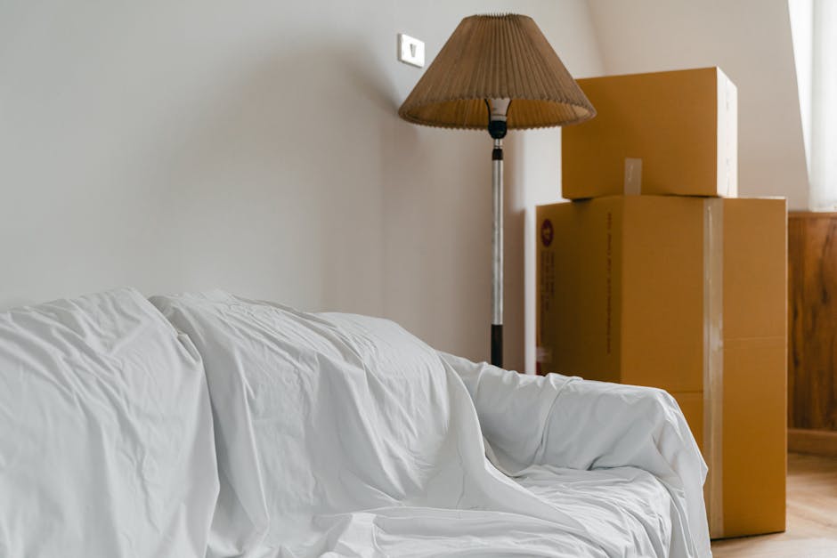 Inside a room during a house relocation, three large cardboard moving boxes are stacked near a wooden cabinet, placed adjacent to a wall with a white electrical outlet visible. A tall, slim floor lamp