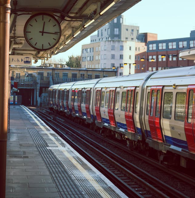 An outdoor view of Enfield Town Station platform showing a wide concrete paved area with a tactile yellow strip along the edge of the platform for safety. The platform is sheltered by a large metal an
