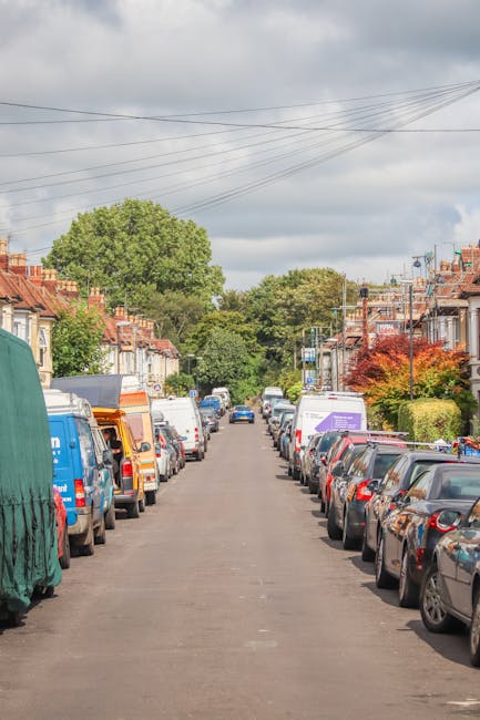 A residential street lined with closely spaced terraced houses, with their front gardens partly visible and some featuring hedges and trees. Numerous parked cars, including sedans, hatchbacks, and van